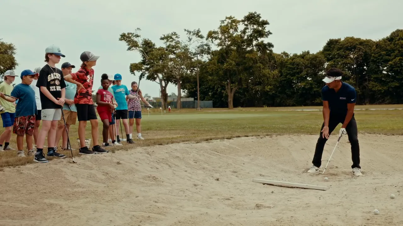 A coach demonstrates a bunker shot while a line of juniors watches