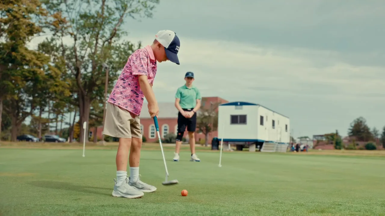 A junior golfer in a pink shirt putts toward the hole while his coach watches